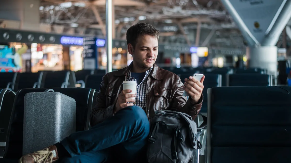 Traveler sitting at an airport gate, checking his phone with a coffee in hand